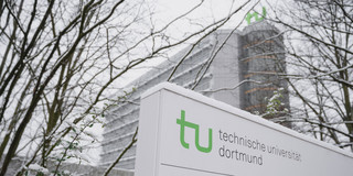 Ein mit Schnee bedecktes Schild mit TU-Logo und im Hintergrund steht das Mathematikgebäude.